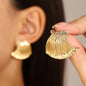 Gold leaf-shaped earring held by a hand with an ear wearing a similar earring in the background.