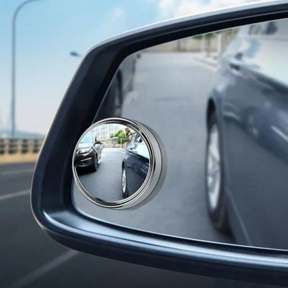 Car side mirror with a small round mirror attached, reflecting another car on a road.