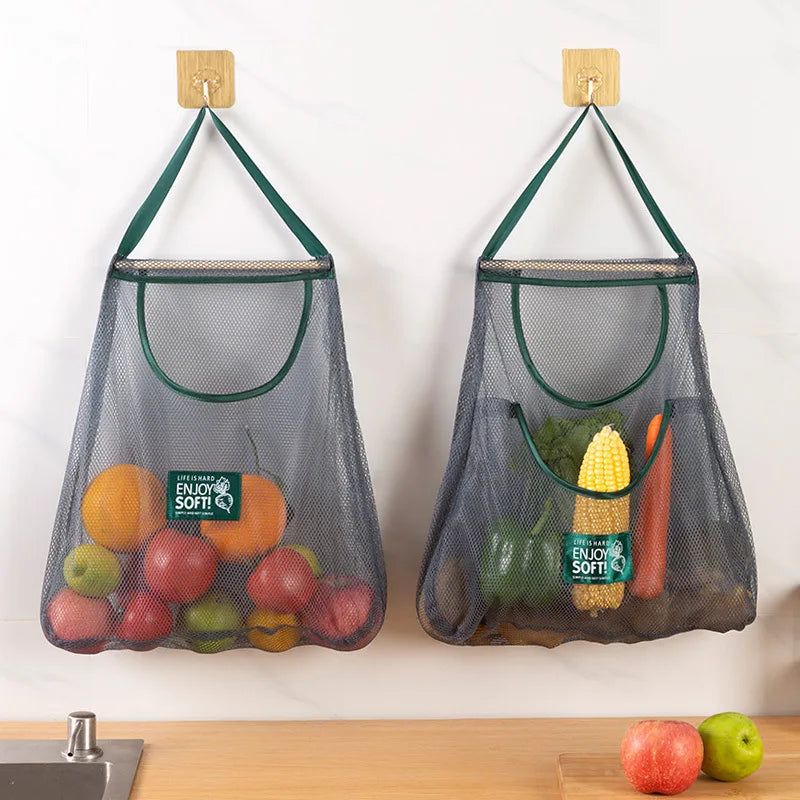 Two mesh grocery bags filled with fruits and vegetables hanging on hooks against a white wall.