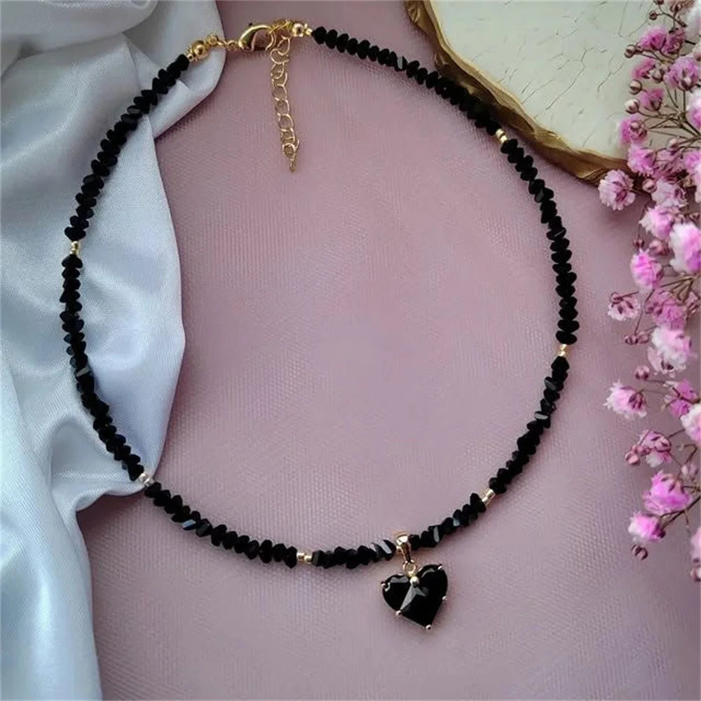 Black beaded necklace with a heart pendant on a person wearing a sheer top, with pink flowers in the background.