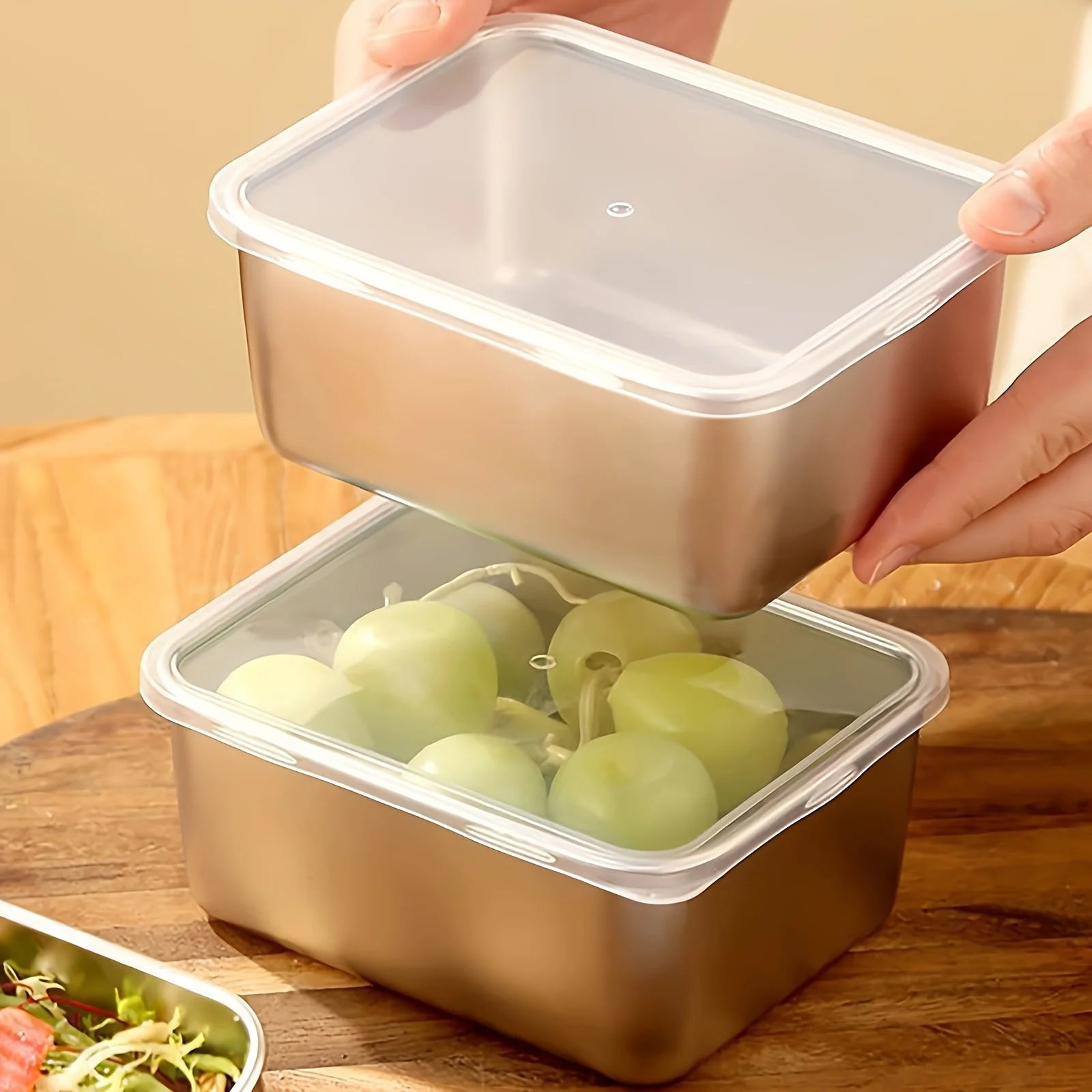 Person holding a metallic food container with green vegetables inside on a wooden surface.