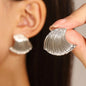 Silver shell-shaped earrings being held up to a person's ear.