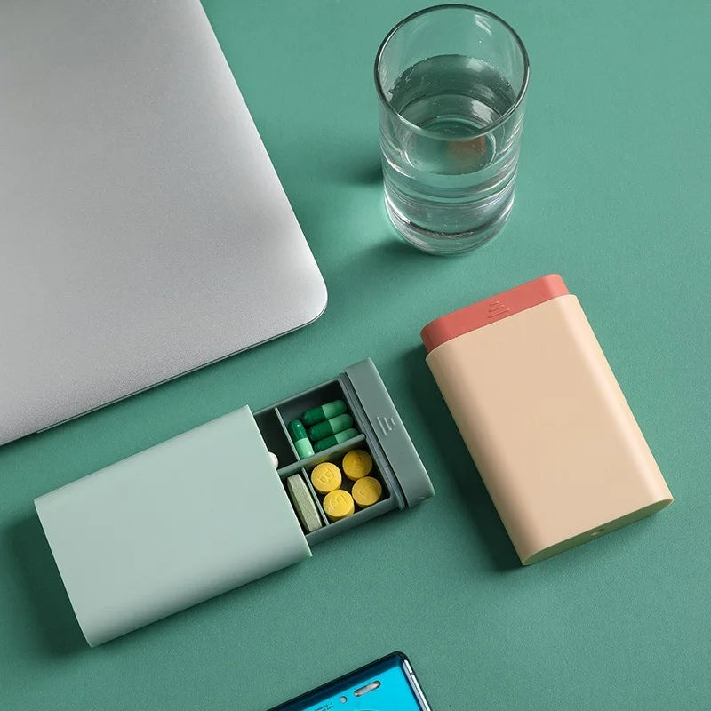 Pill organizer with medications on a green surface next to a glass of water, phone, and laptop.
