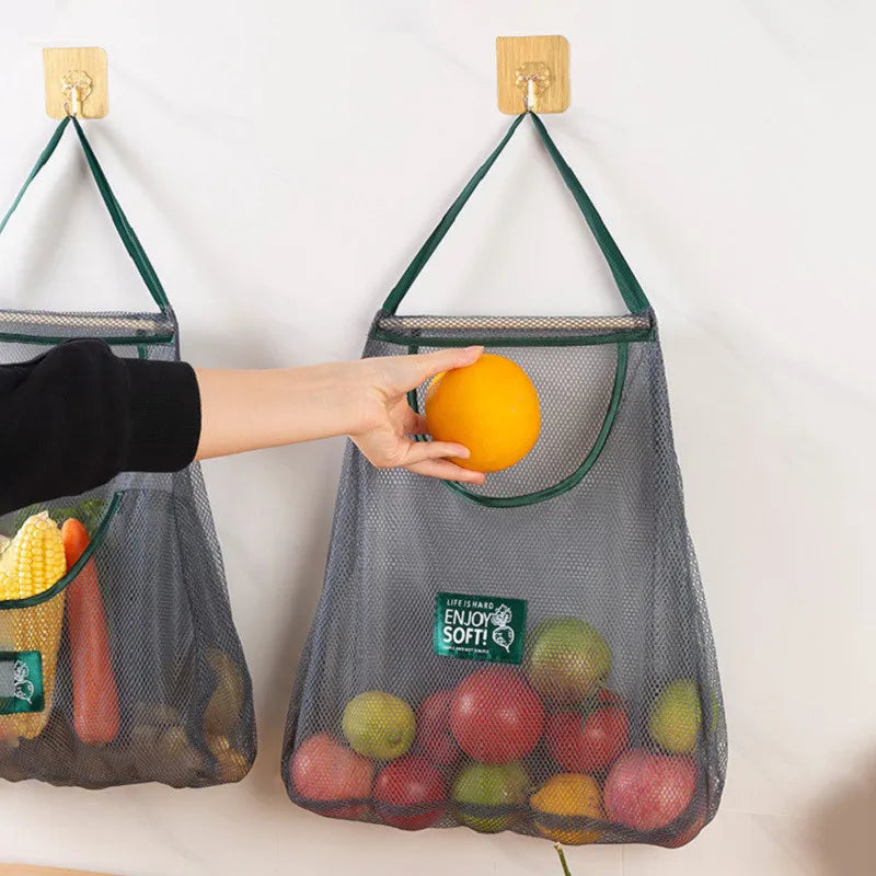 Mesh grocery bags with fruits and vegetables hanging on hooks against a white wall.