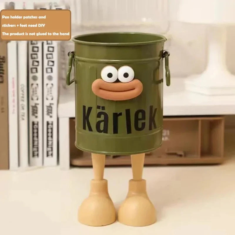 Green bucket with a face and 'Kärlek' text, standing on a surface with books in the background.