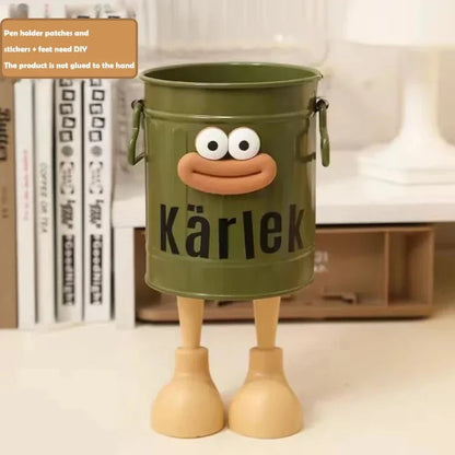 Green bucket with a face and 'Kärlek' text, standing on a surface with books in the background.